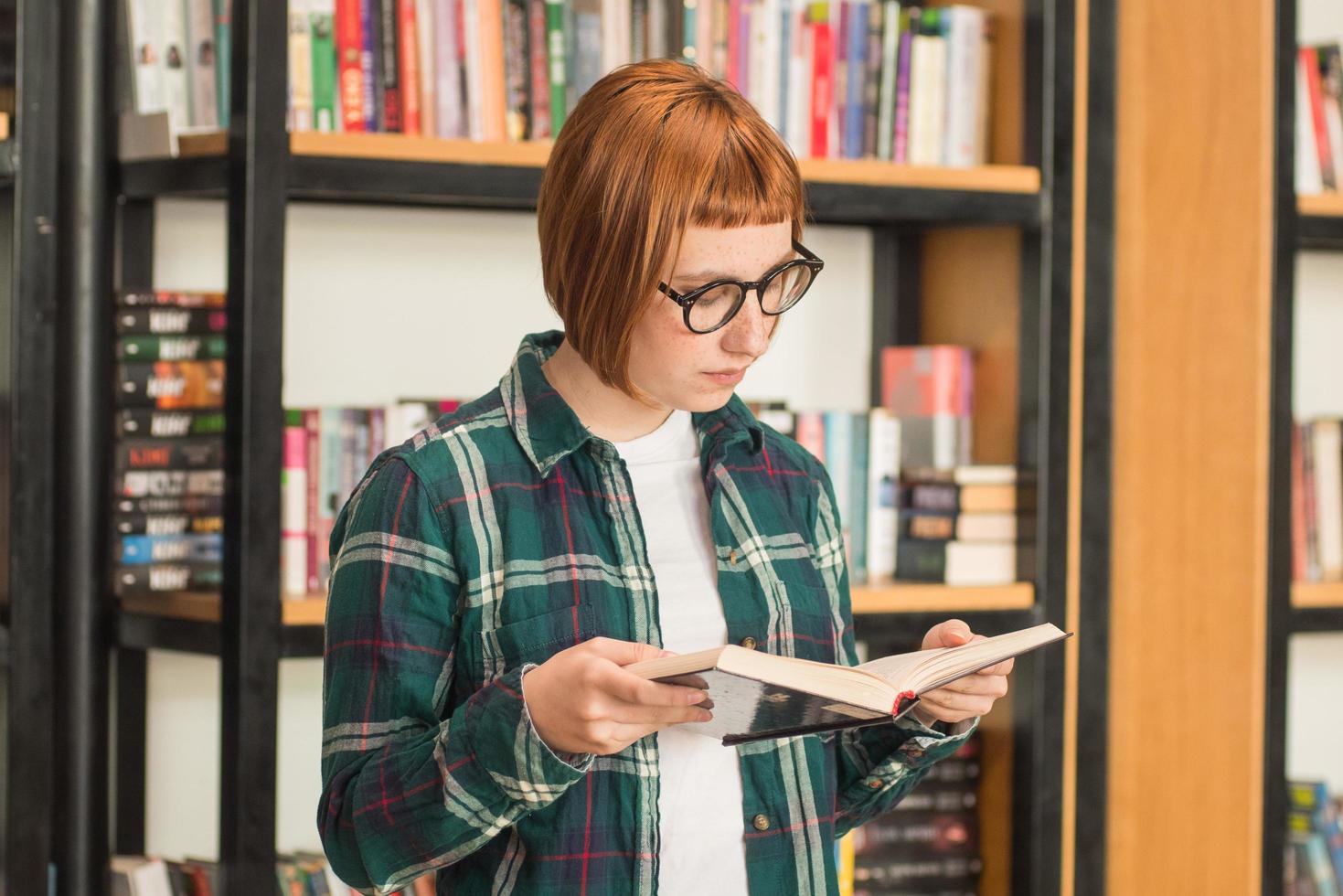 joven pelirroja con gafas lee un libro en la biblioteca foto