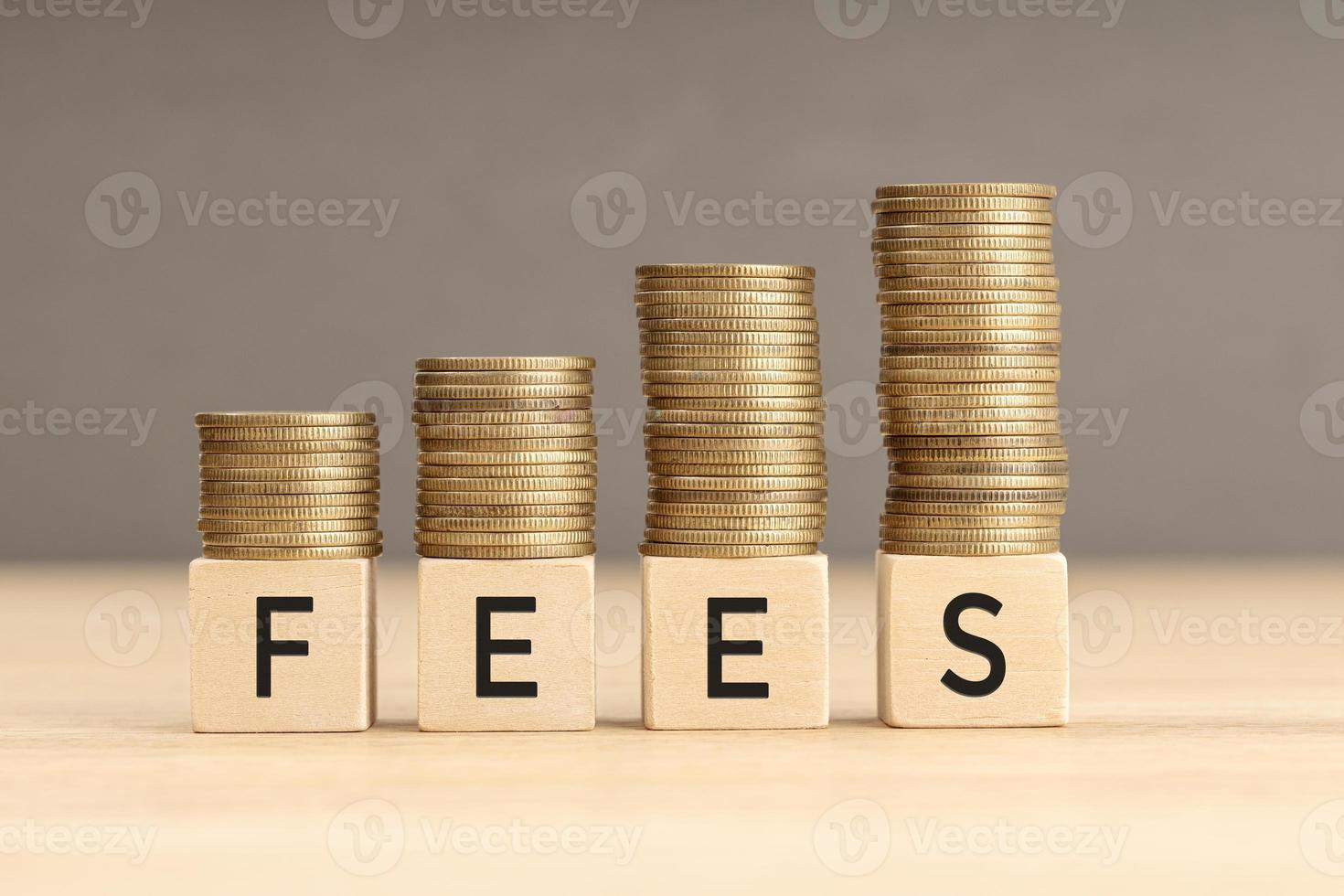 Fees word in wooden blocks with coins stacked in increasing stacks. Fees increasing concept. Copy space photo