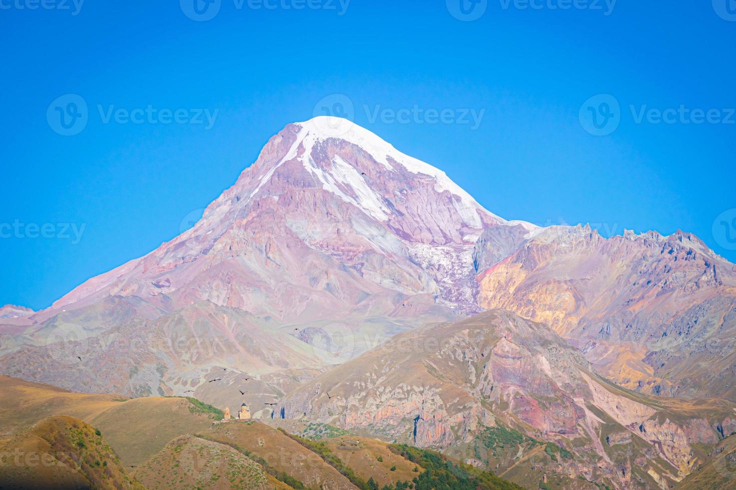 Beautiful blue panorama of gergeti trinity church with stunning view of Kazbek peak in background photo