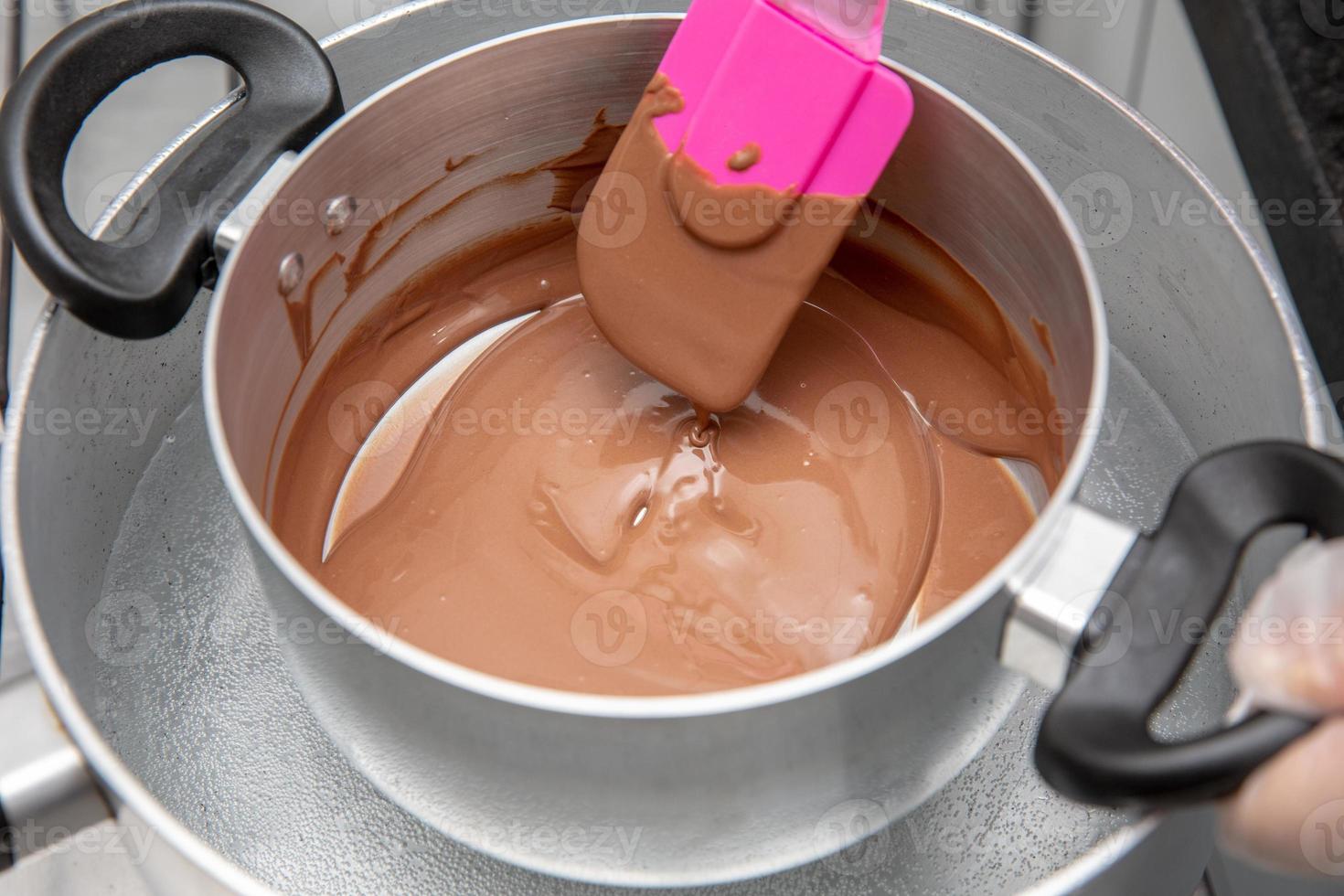 Cooking pot with chocolate melting being removed by chef hand on the stove of a kitchen to make sweet desserts. Bulk chocolate melting in a pot. photo