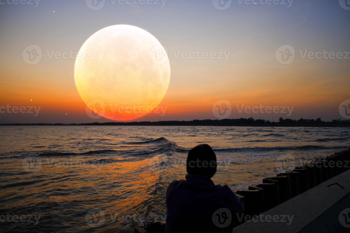 hombre en silueta mirando a la distancia la tranquila luna llena naranja en el extranjero con un terciopelo púrpura y un cielo naranja tranquilo. foto