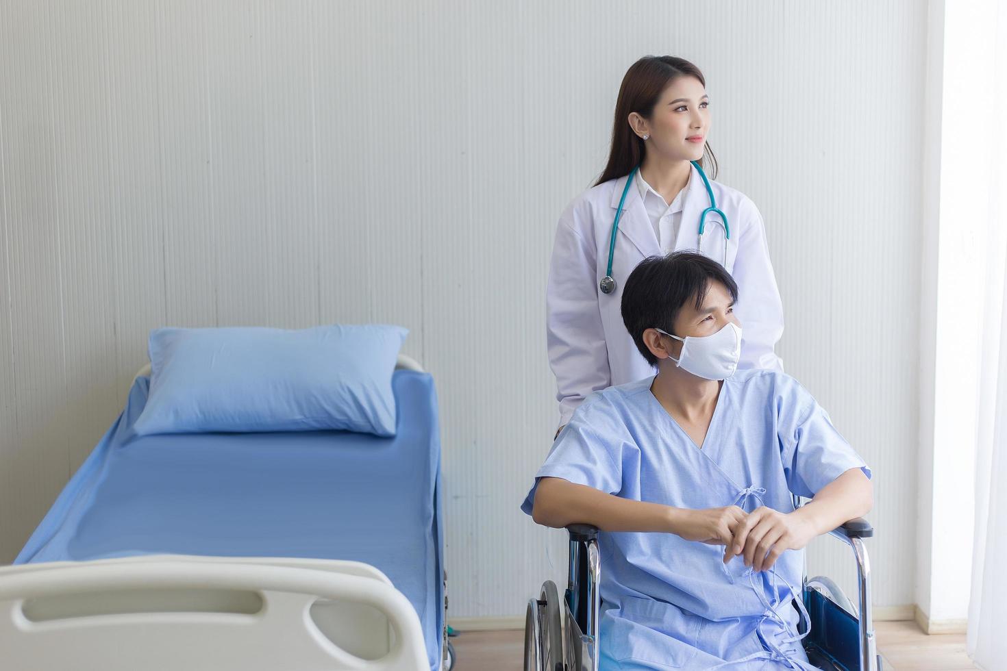 Asian doctor woman talking with a man patient who wear face mask about his health symptom while he sit on wheelchair at hospital. photo