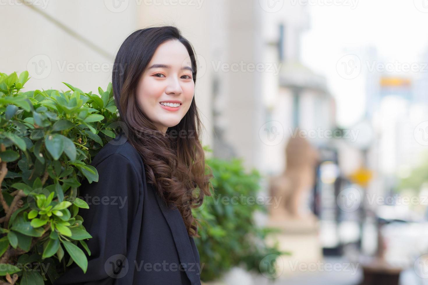 un retrato de una mujer asiática de pelo largo con una túnica azul oscuro sonriendo y felizmente parada al aire libre en la ciudad. foto