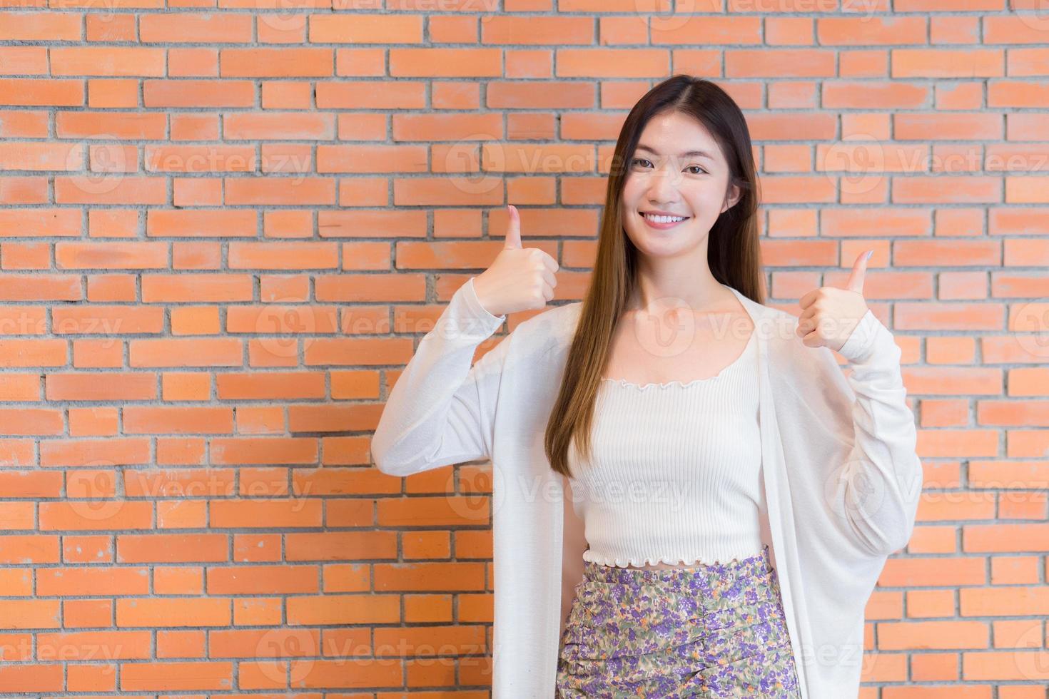 Beautiful business young Asian woman who has blonde long hair with a white shirt smiles and shows thumb up and presents something on a brown brick background. photo