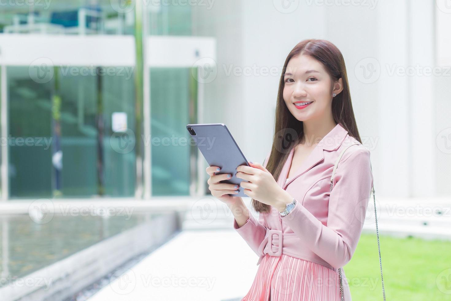 Beautiful young Asian professional business woman with long hair is smiling outdoor in the garden front of the office while hold at the tablet in her hand to go out meeting with clients at the office. photo