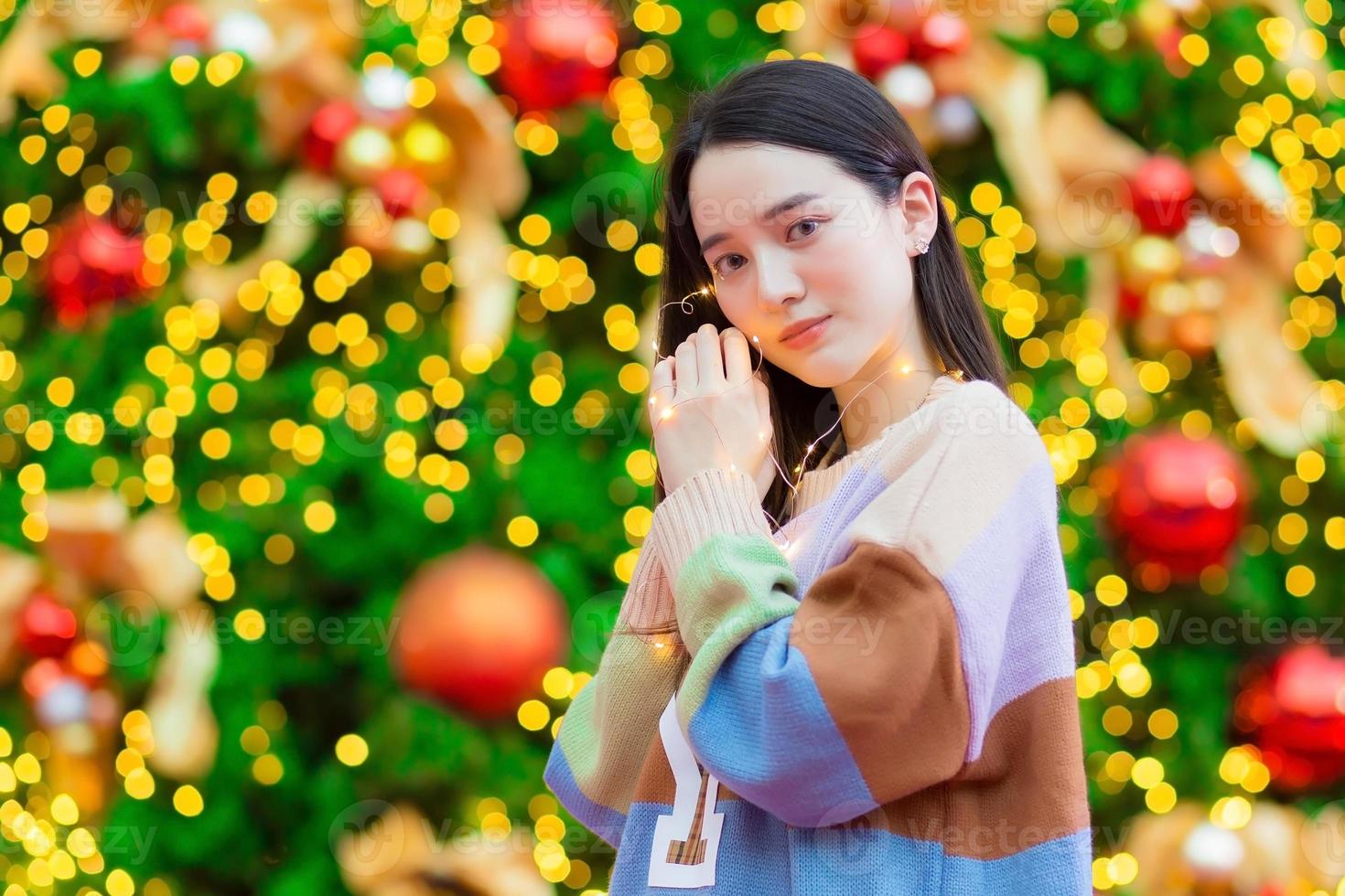 Asian beautiful woman in a colorful sweater stands happily. In his hand he held a light in front of the Christmas tree. With bokeh as background In the theme of Christmas and New Year celebrations photo