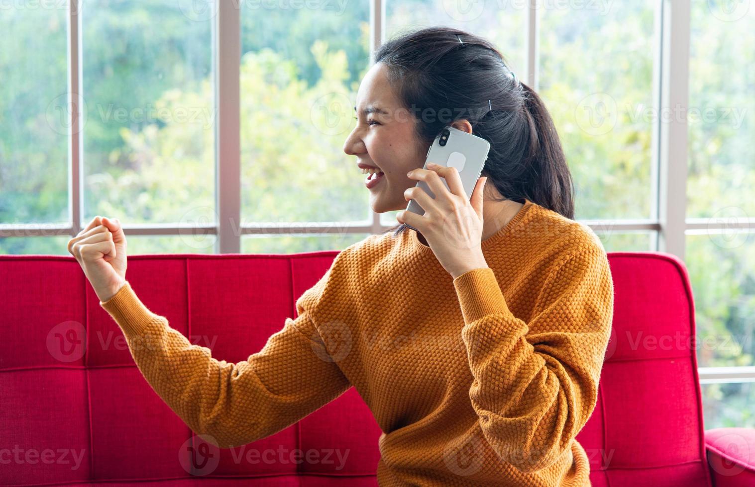 atractiva mujer hermosa con teléfono inteligente sentada en el interior de un sofá rojo, haciendo una llamada telefónica con una emoción emocionante foto