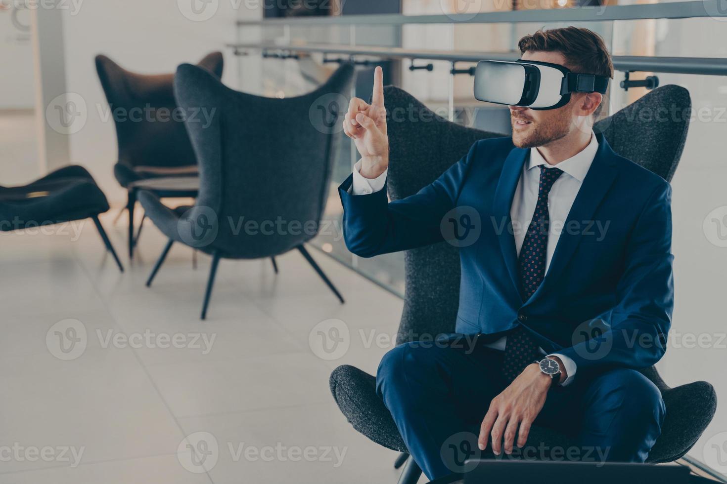 Young CEO sitting in office centre lobby with VR headset goggles, pointing with forefinger up in air photo