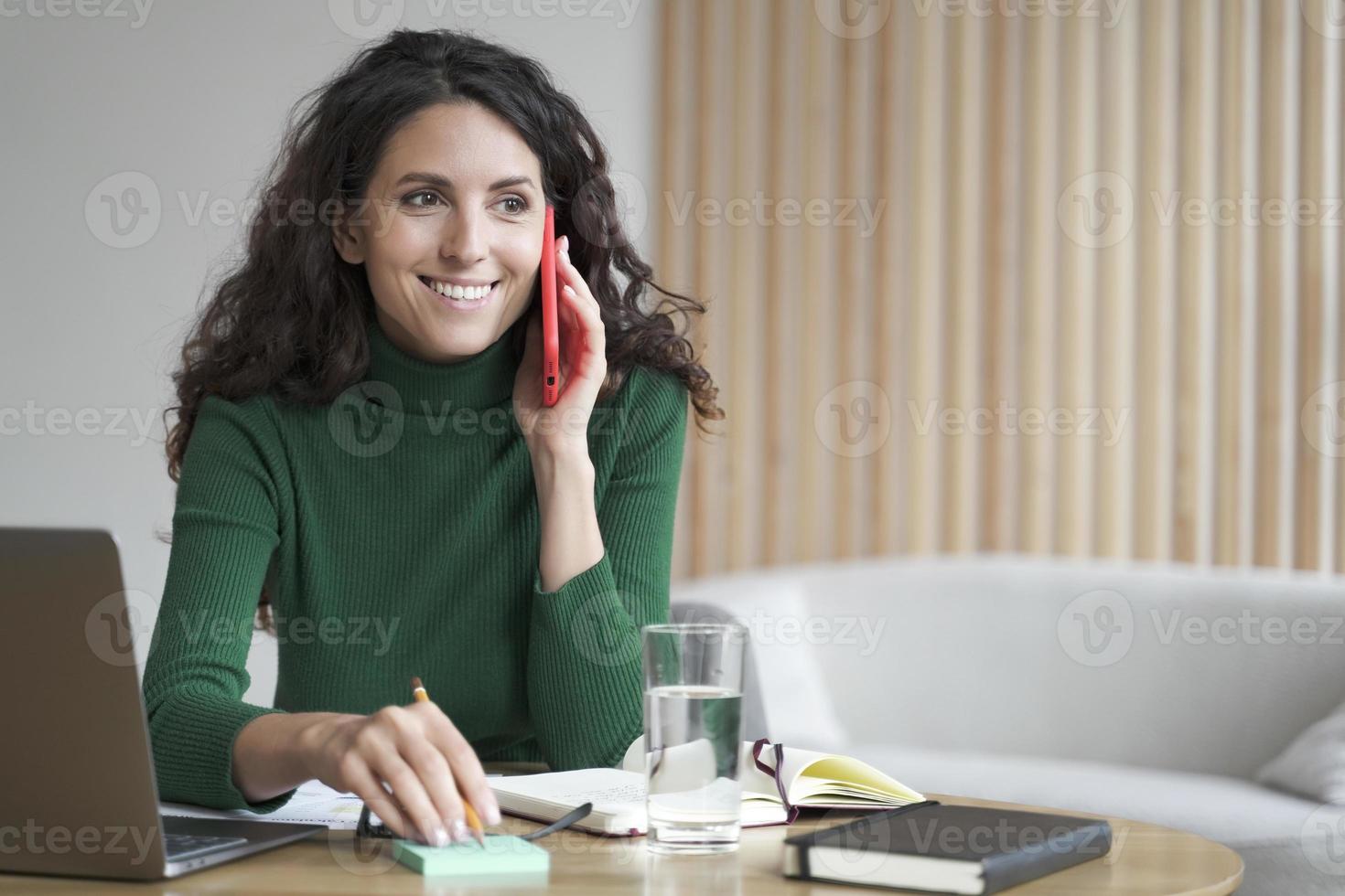 Smiling business lady using mobile to communicate and receive orders, writing info on sticky note photo