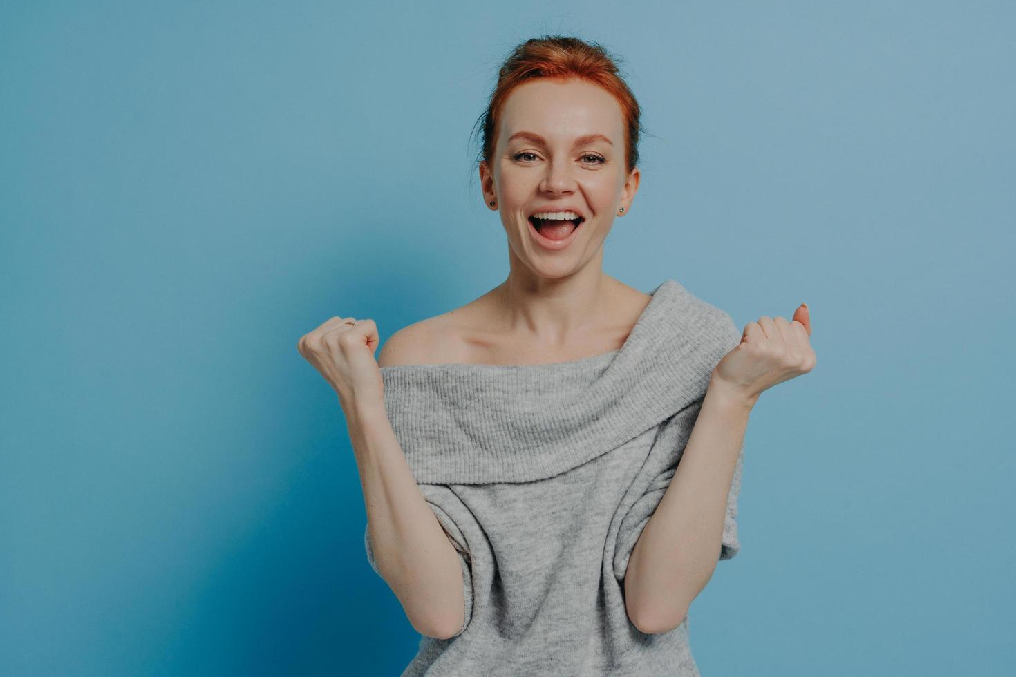 Young overjoyed excited red haired woman sweater clenching fists with triumph photo