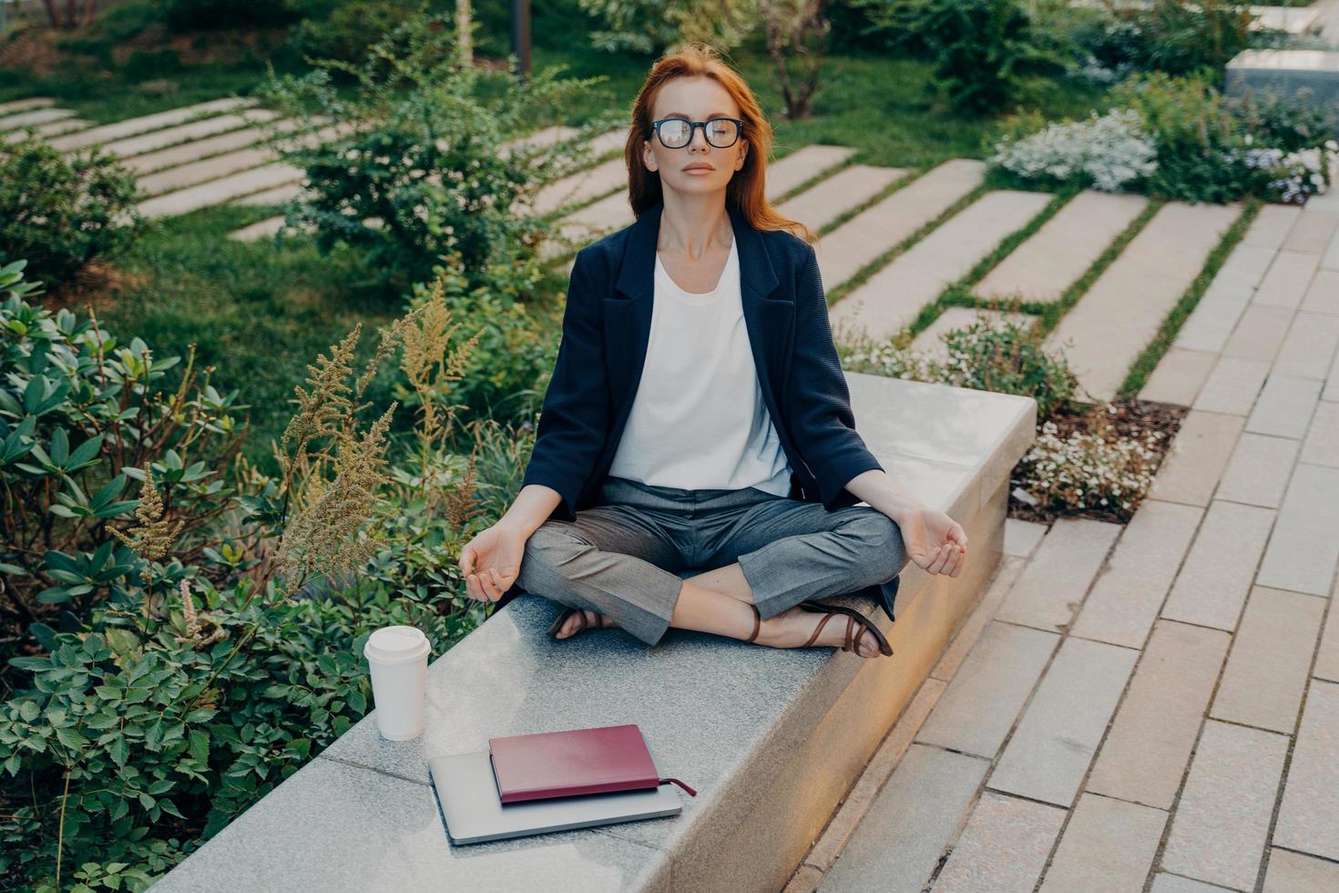 Redhead young European woman sits in lotus position wears formal clothes drinks takeaway coffee photo