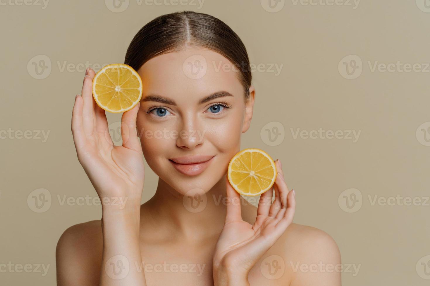 Portrait of young brunette woman with dark combed hair looks directly at camera holds two slices of lemons stands topless has healthy glowing skin isolated over beige background. Facial care concept photo
