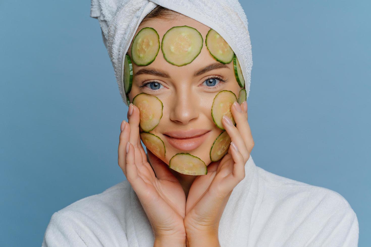 Close up shot of young woman applies cucumber slices on face for healthy skin looks directly at camera dressed in bath dressing gown and towel on head isolated on blue background. Facial care concept photo