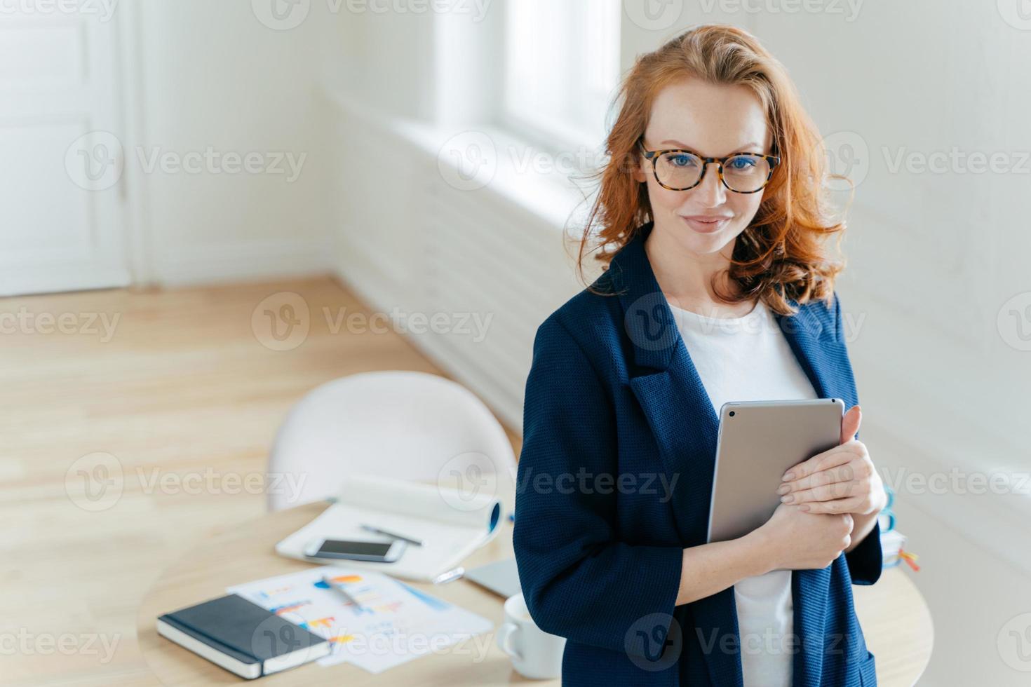 una foto de una hermosa joven pelirroja revisa la notificación en el panel táctil, feliz de recibir un mensaje de ingresos, mira con confianza a la cámara, el lugar de trabajo con documentos en papel y el bloc de notas en segundo plano.