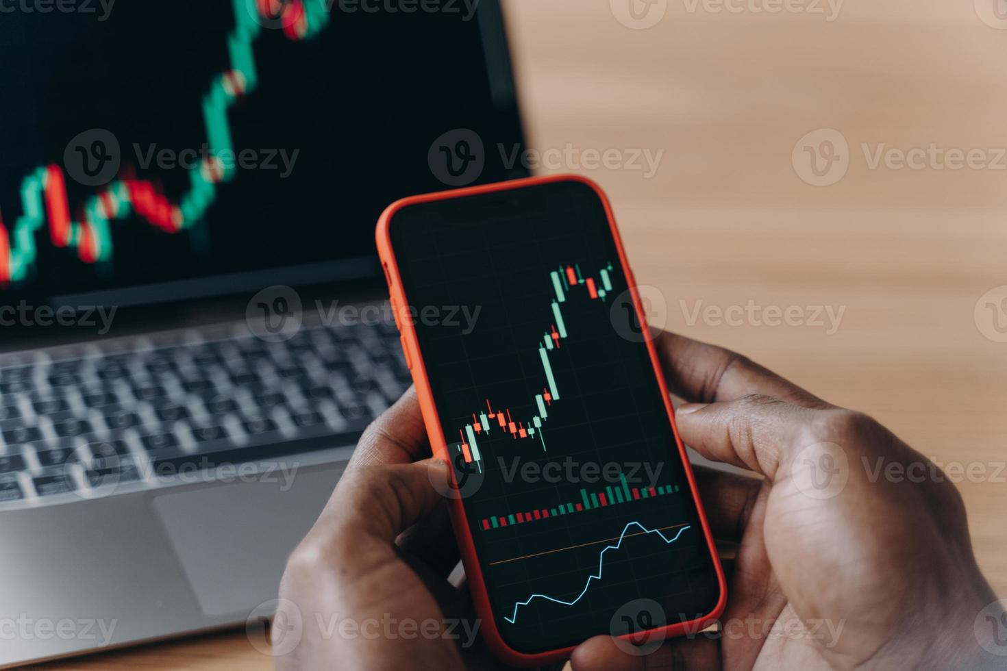 Male hands of African man holds smartphone in front of laptop with trading charts on it photo