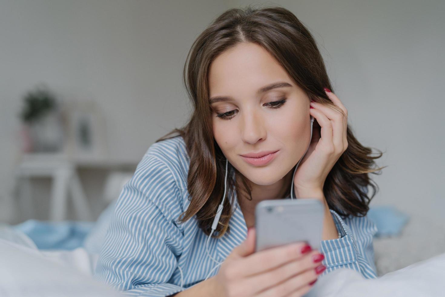 una foto de una guapa estudiante morena aprende el idioma a través de una aplicación móvil, escucha audio con auriculares, tiene tiempo libre, se viste con ropa informal, posa en una cómoda cama blanca, tiene una mirada pensativa a un lado