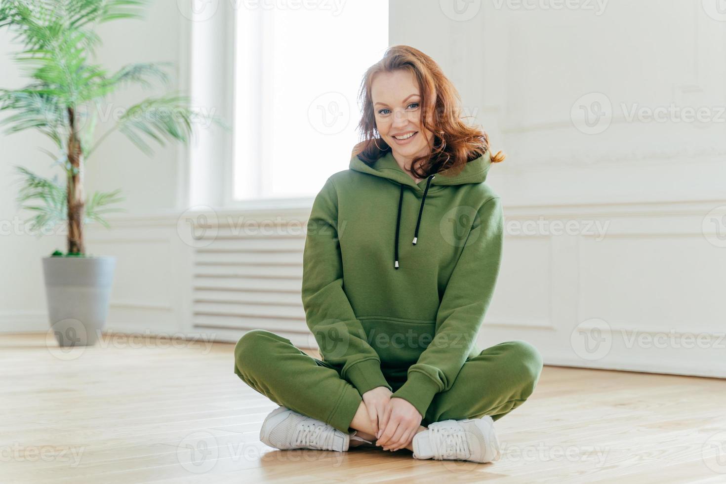 Happy ginger female sport trainer sits crossed legs on floor, wears green hoody and trousers, white trainers, smiles gladfully, poses on floor in empty room for going in for sport. Healthy lifestyle photo