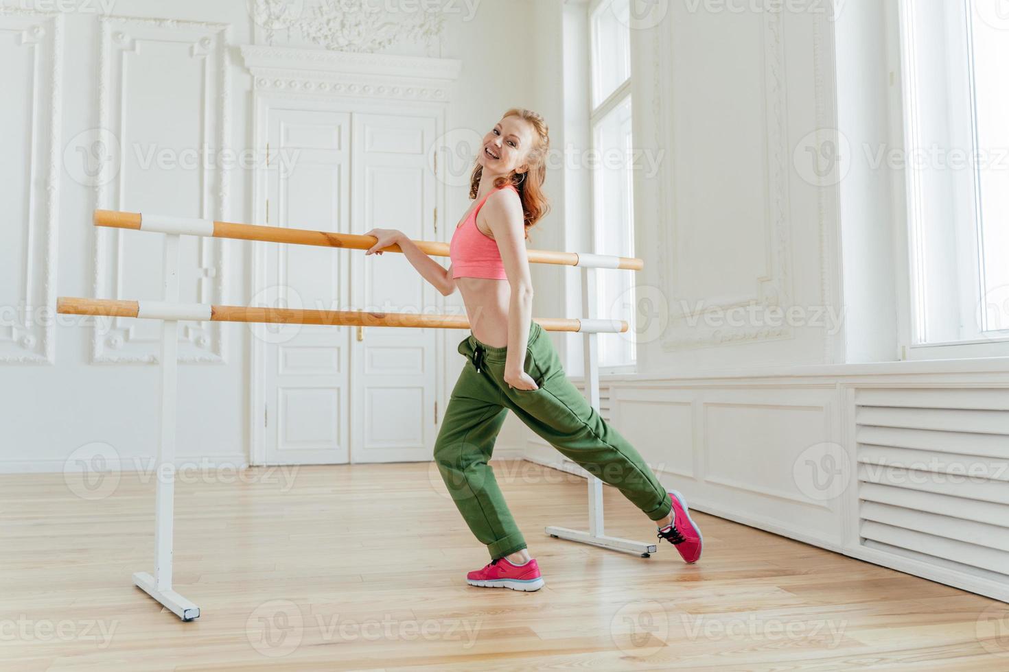 Motivated cheerful red haired female in sports bra and trousers, keeps hand in pocket, wears sneakers, has exercises on ballet barre, dances at studio, being in good mood, finishes dance workout photo
