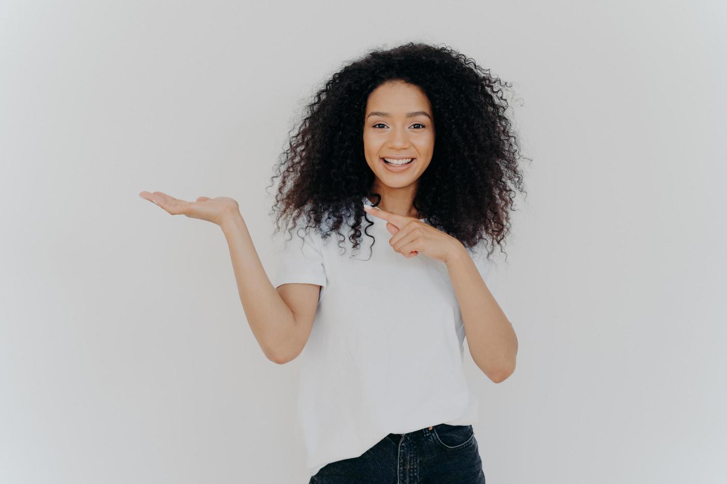 Choice is simple. Attractive feminine girl with Afro hair raises palm and points on blank space, makes decision, has toothy smile, wears casual wear, poses against white background. Look at this photo