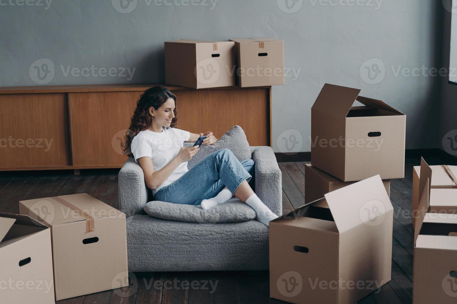 Woman in armchair clicks phone among boxes. European girl having rest in apartment after relocation. photo