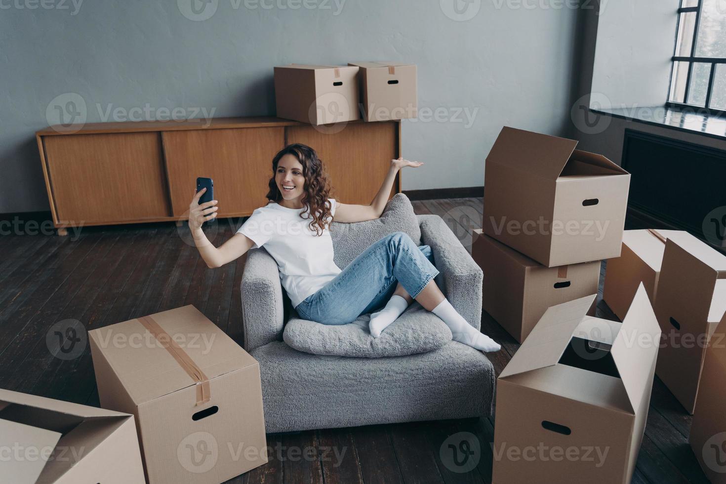 Happy spanish girl sitting in armchair and having video call on smartphone. Owner in new apartment. photo