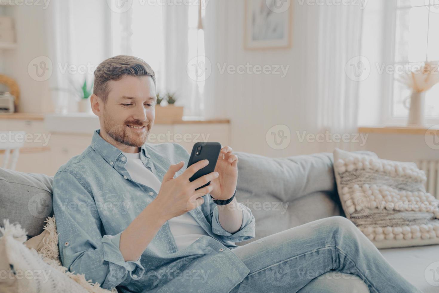 hombre barbudo sonriente feliz usando un dispositivo de teléfono inteligente mientras se relaja en el sofá foto