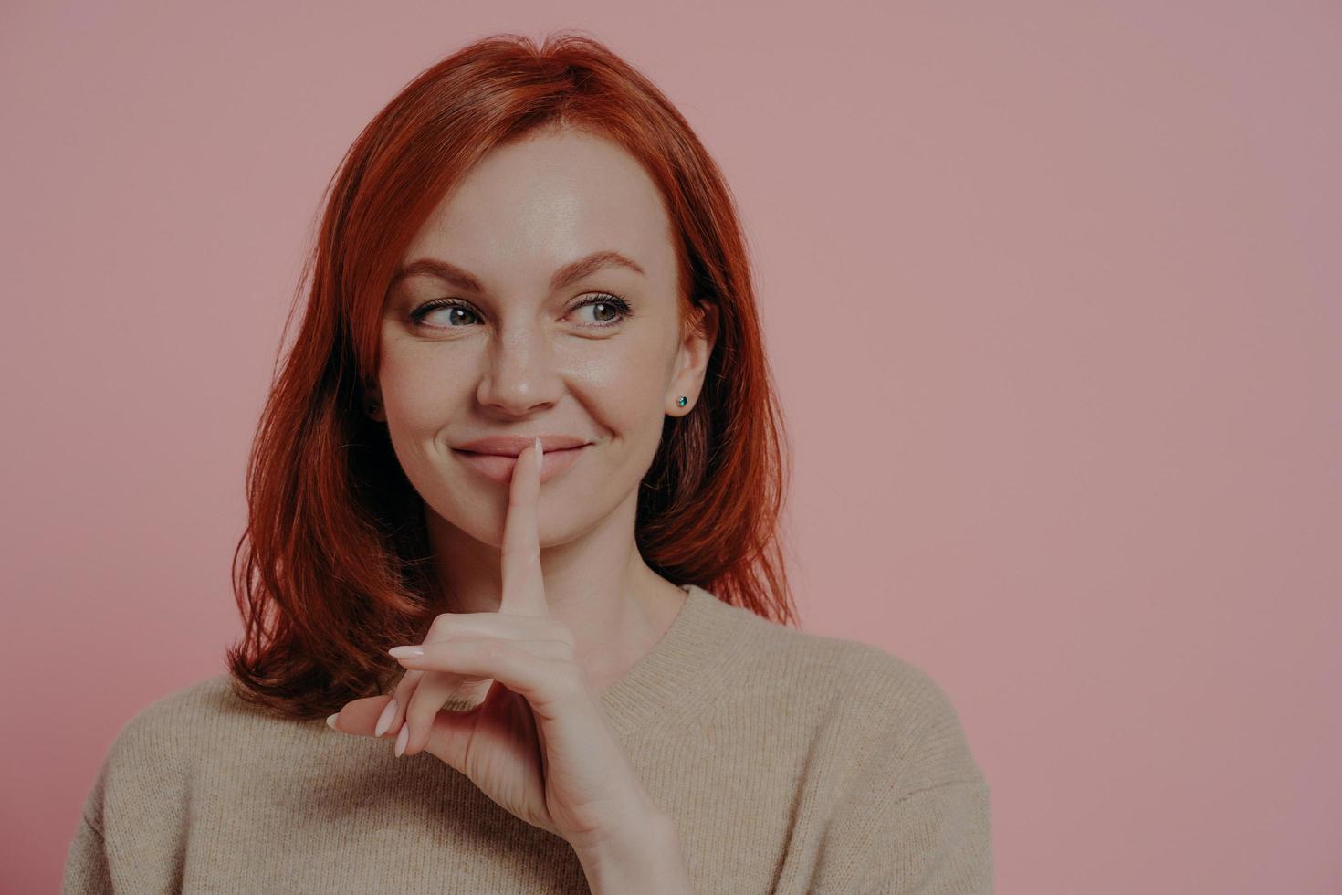 hermosa joven pelirroja sonriente poniendo el dedo en los labios y haciendo el signo de silencio foto