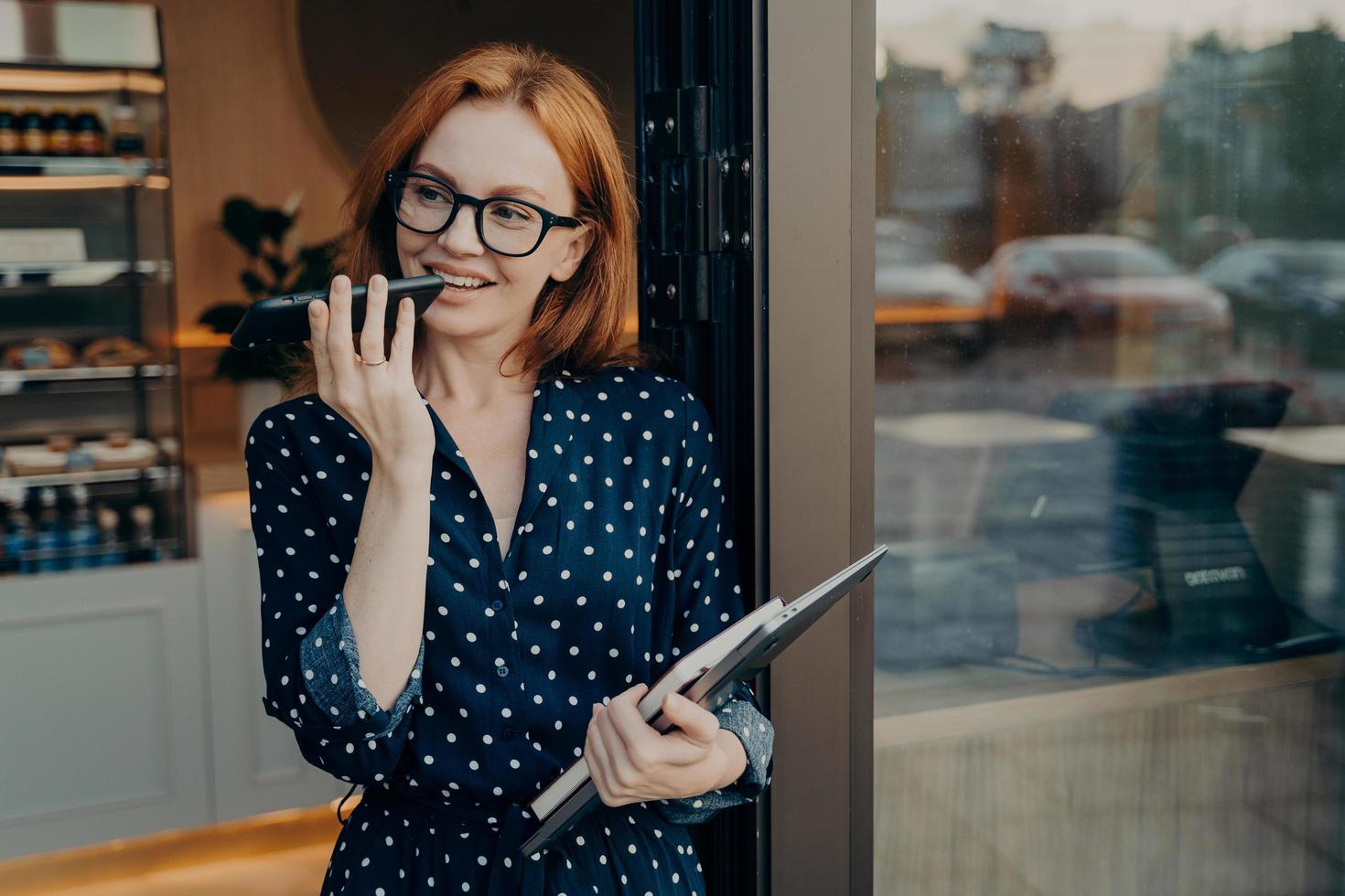 Female freelancer stands outdoors with mobile phone and uses voice recognition system on smartphone photo