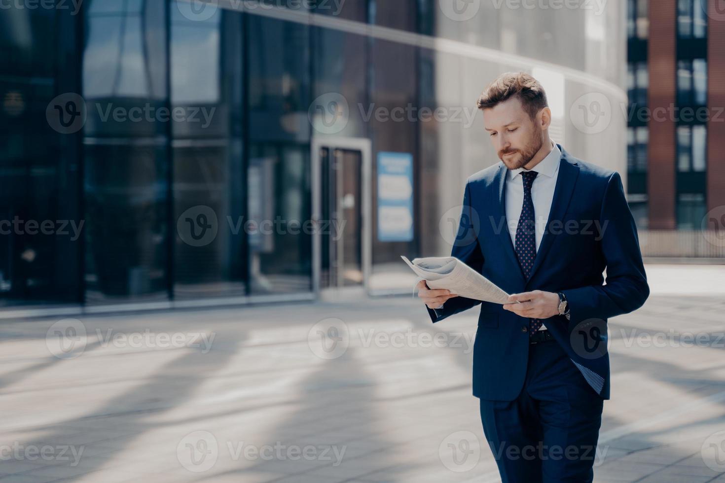 Focused formally dressed business person reading newspaper while walking near office building photo