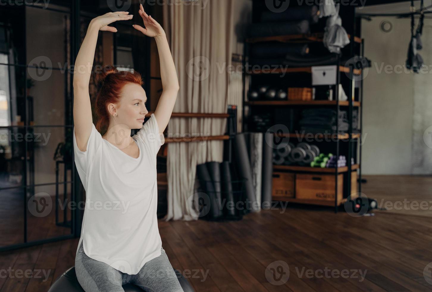 Sportive young woman with red hair sits on big pilates ball with hands lifted over head photo