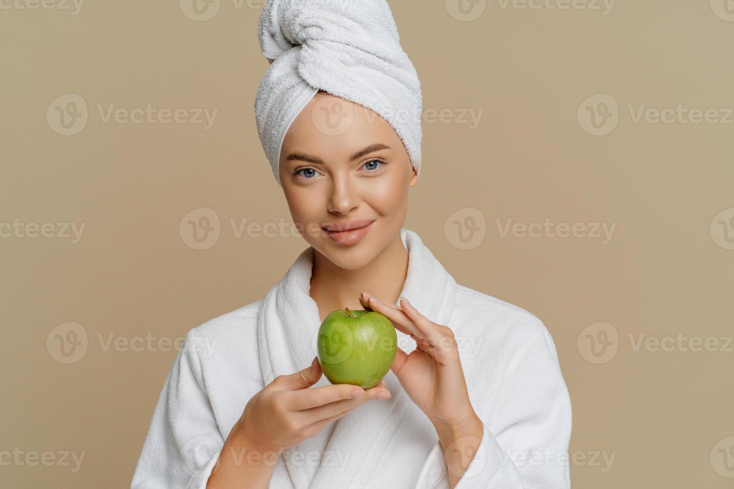 Lovely young woman has refreshed skin after taking bath dressed in dressing gown wrapped towel on head holds green apple containing much vitamins isolated over brown background. Hygiene concept photo