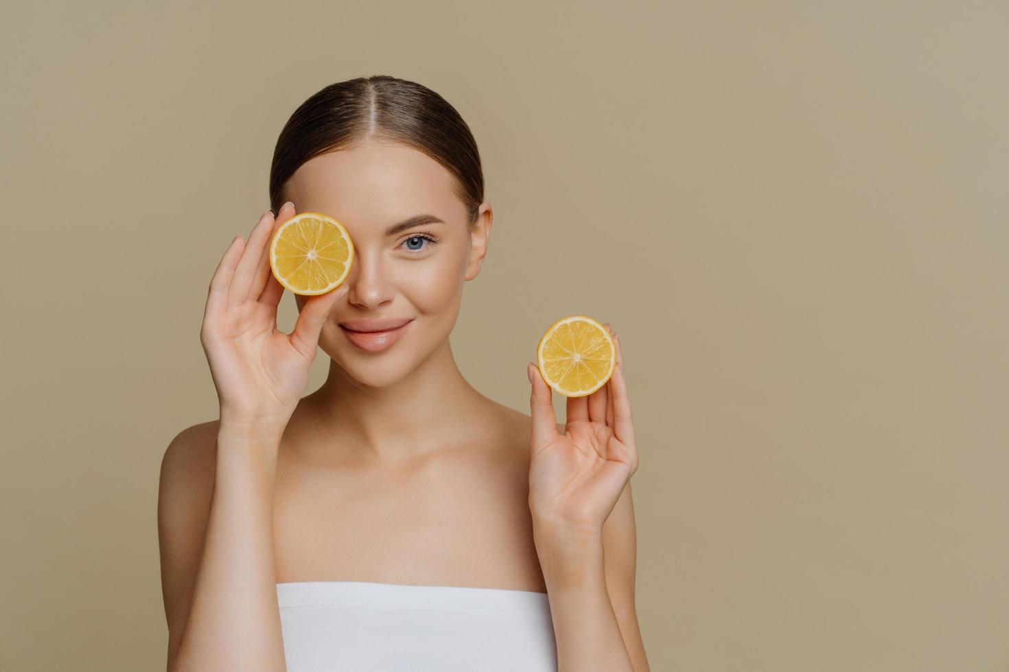 limón para la belleza y la salud. hermosa joven saludable cubre los ojos con la mitad de limón disfruta de un tratamiento de spa envuelto en una toalla de baño aislada sobre un espacio de copia de fondo beige para su promoción foto