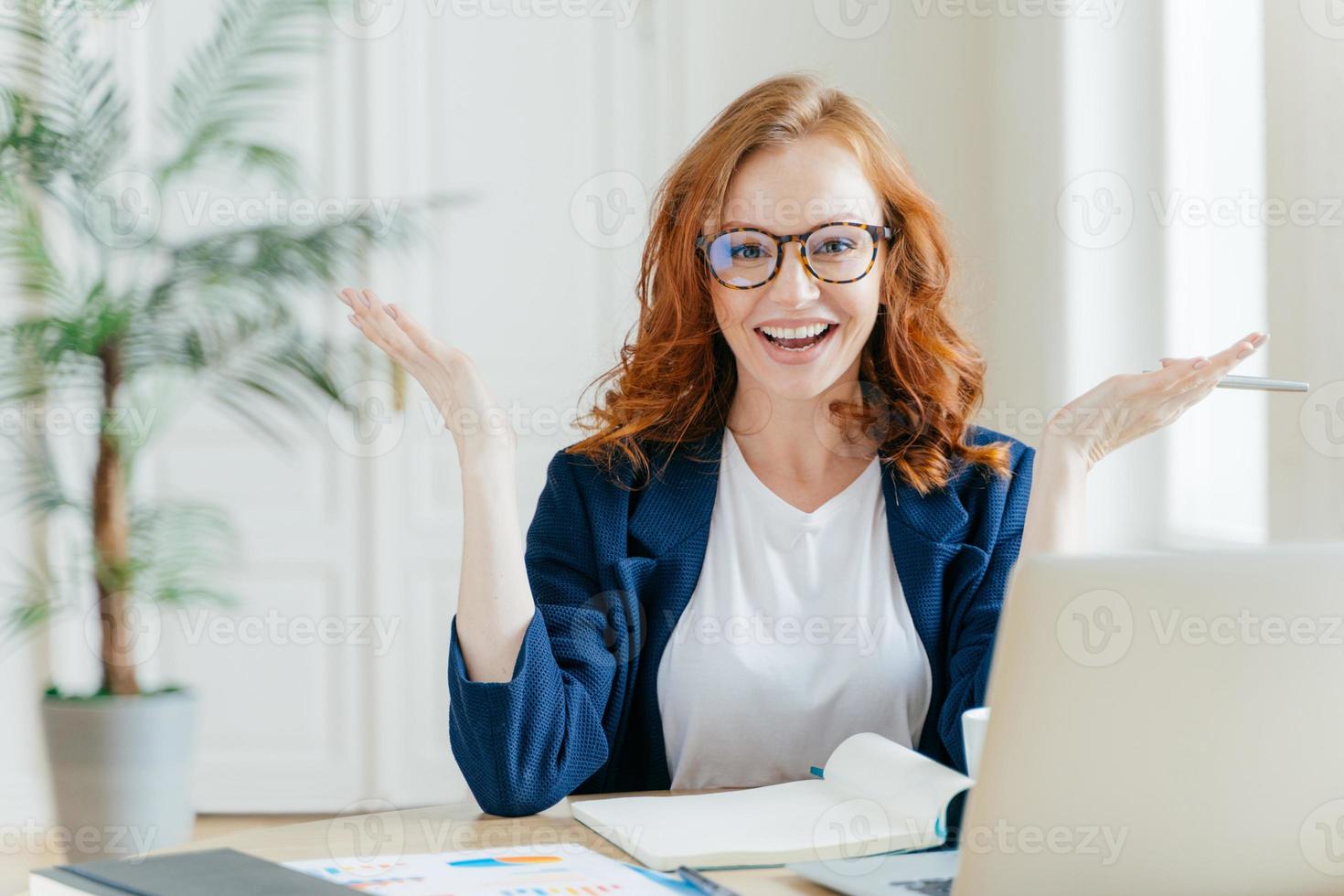 Shot of positive red haired female freelancer works remotely with paper documents, spreads hands to show her good result, sits at desktop with notepad and laptop computer, prepares course work photo