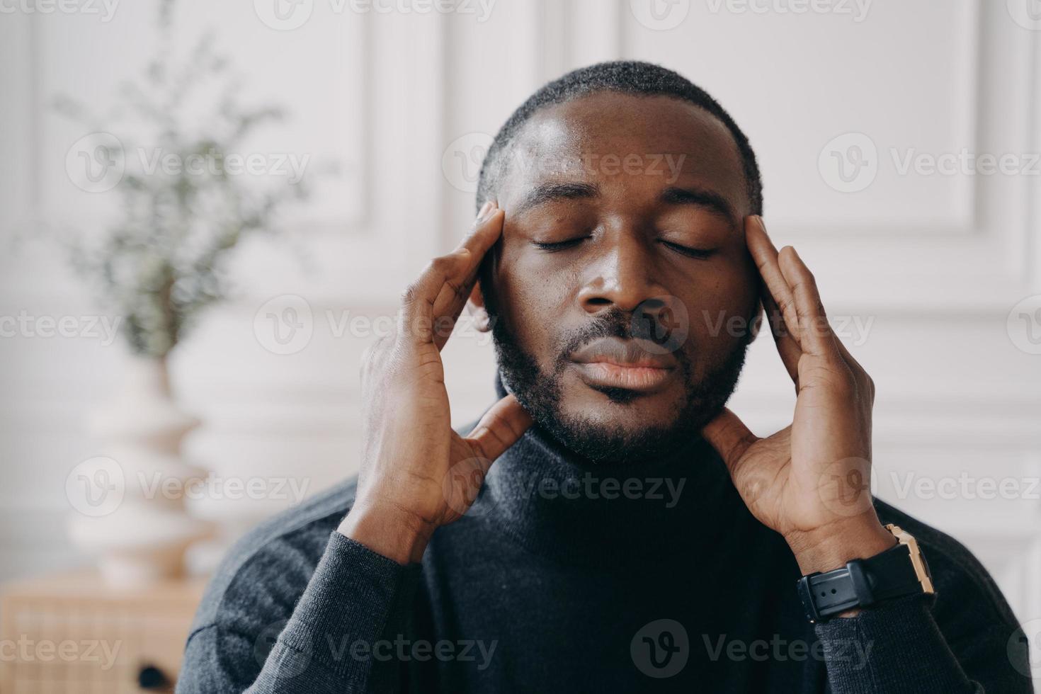 Stressed African home office worker sits at desk touching temples while eyes closed photo