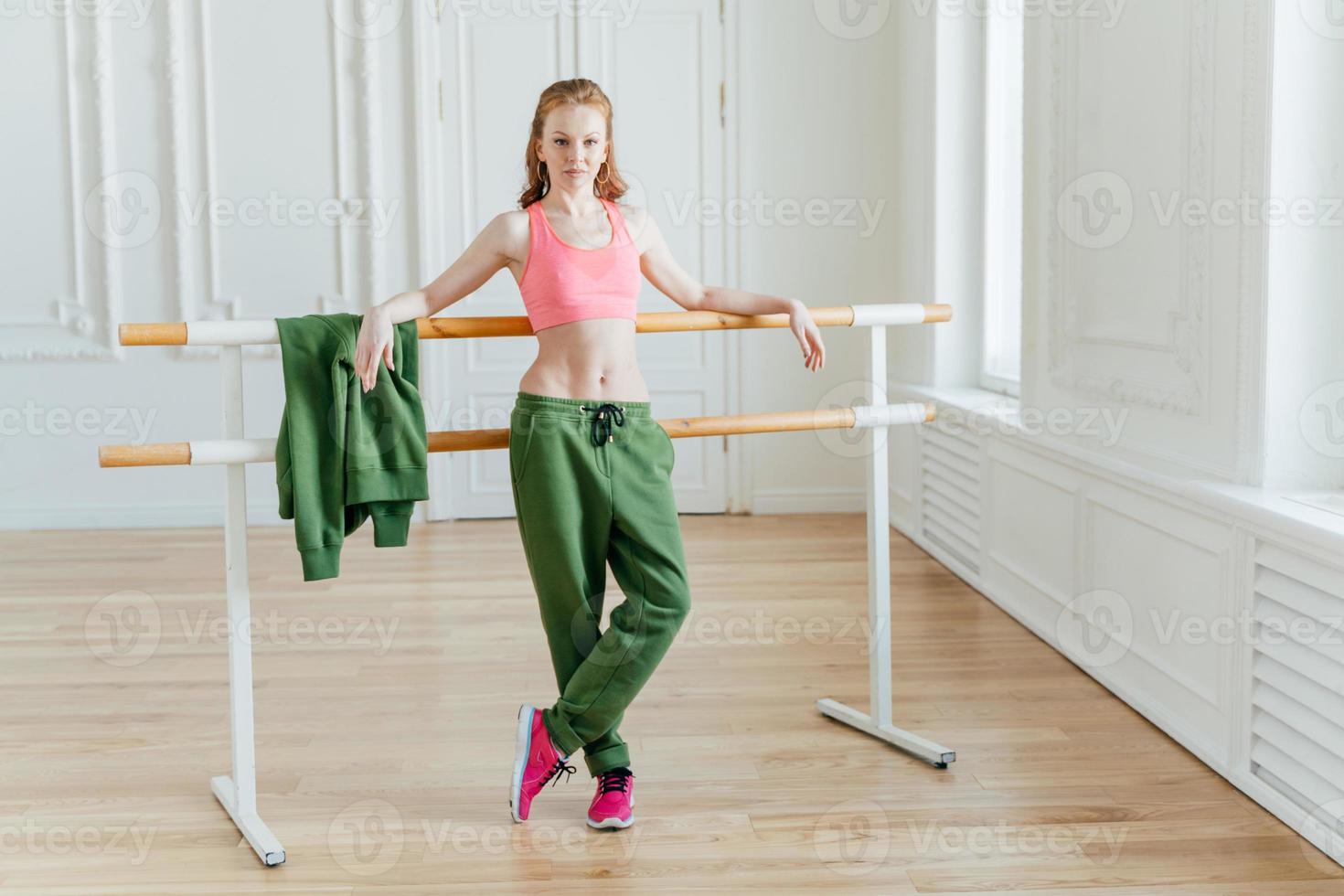 Slim beautiful female ballet dancer leans at ballet barre, wears pink top, sport trousers and sneakers, shows fit figure, stretches before dancing, poses in studio or classroom, has rehearsal photo