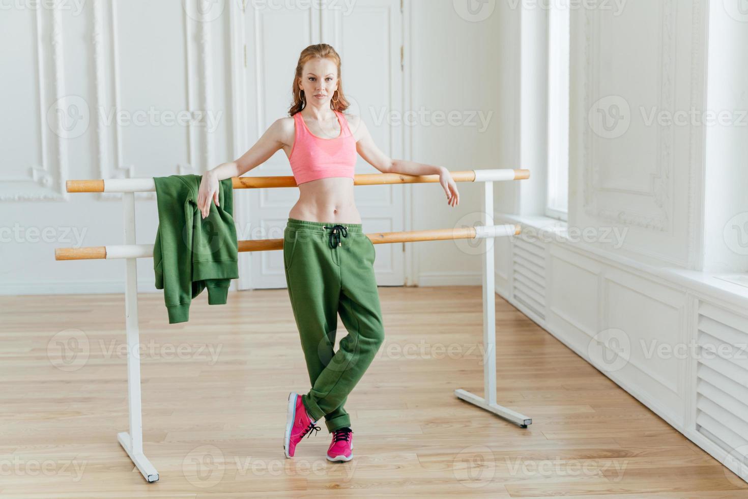 delgada y hermosa bailarina de ballet se inclina en la barra de ballet, usa top rosa, pantalones deportivos y zapatillas de deporte, muestra una figura en forma, se estira antes de bailar, posa en el estudio o en el aula, tiene ensayo foto