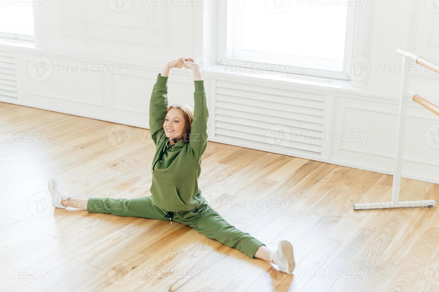 Strong flexible curly red haired woman does side leg split, raises hands, wants to have perfect body, wears sportsclothes, has training in ballet studio. Talented dancer has rehearsal indoor photo