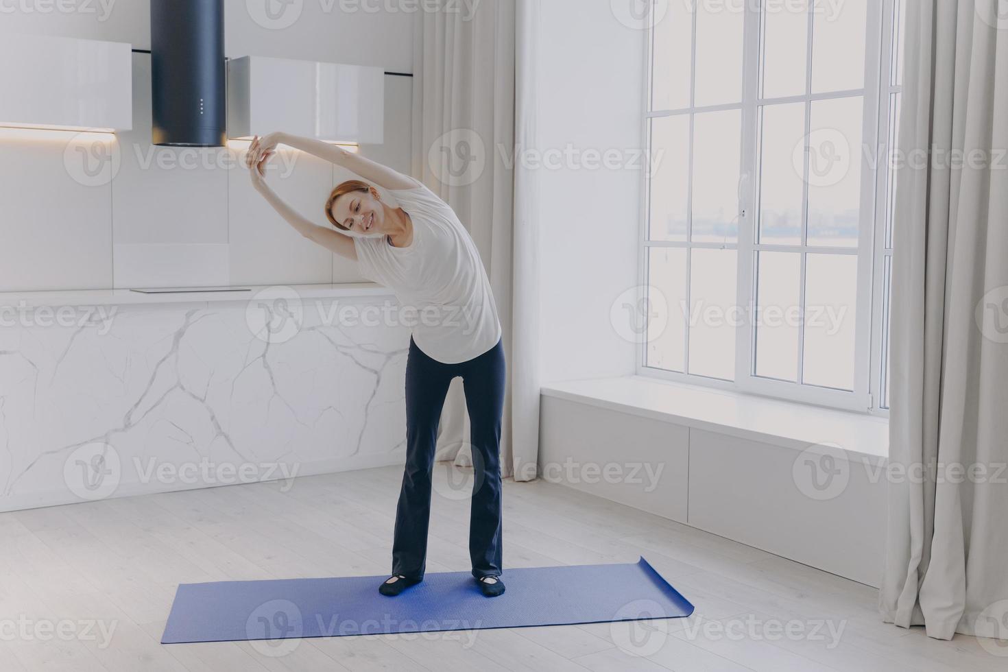 una joven blanca con ropa deportiva y camiseta blanca se estira en casa. Ejercicio de mañana. foto