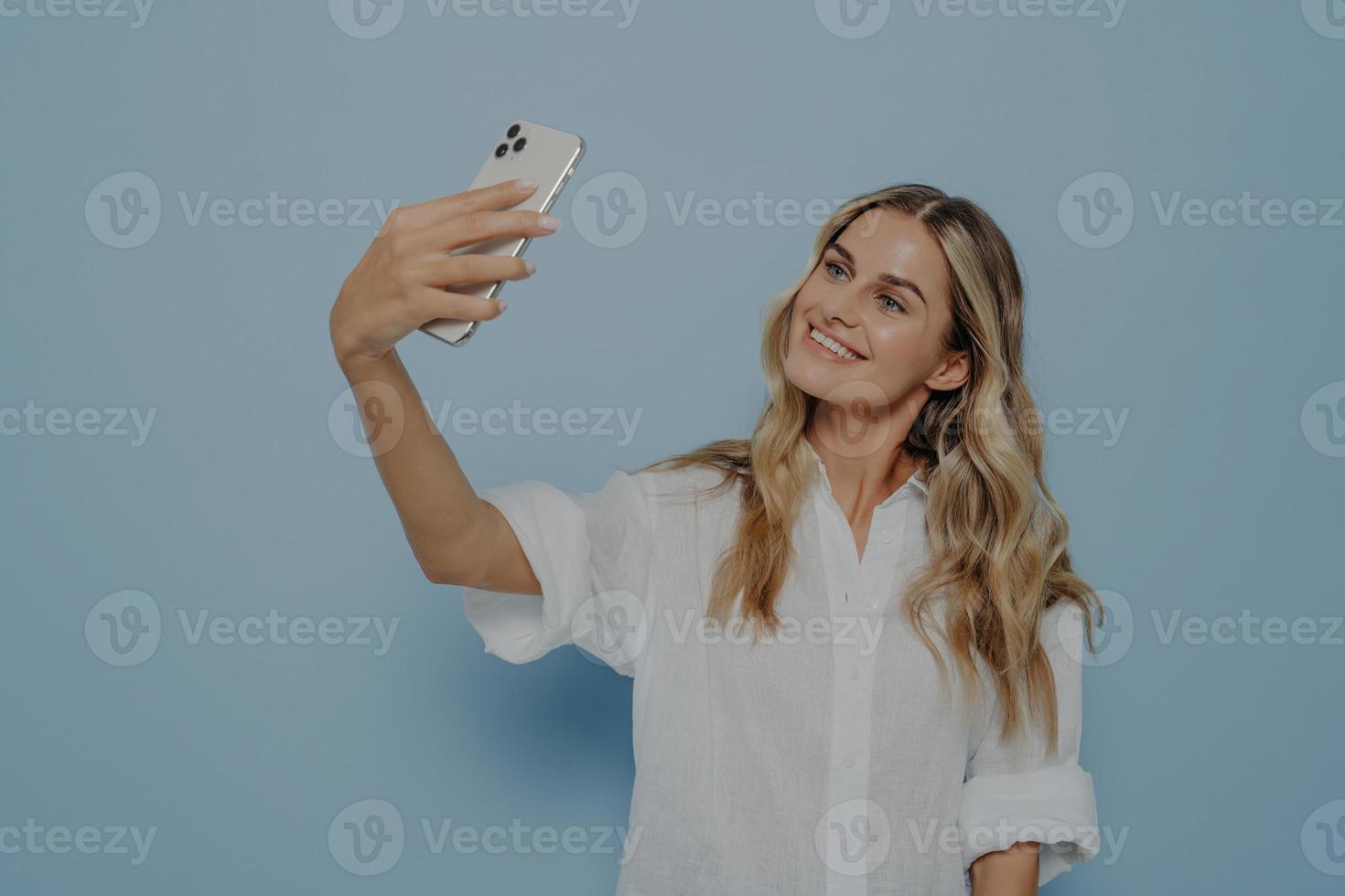 Beautiful young female with long blonde dyed hair holding mobile phone and posing for selfie photo