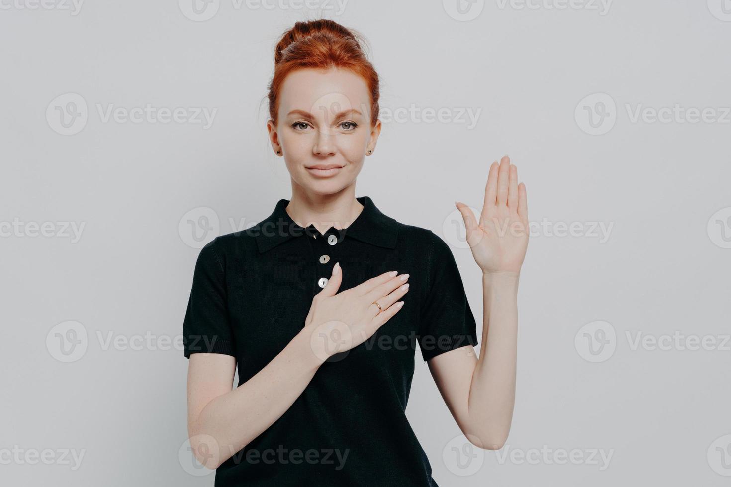 tudio shot of serious red haired female making promise, swearing with hand on chest in studio photo