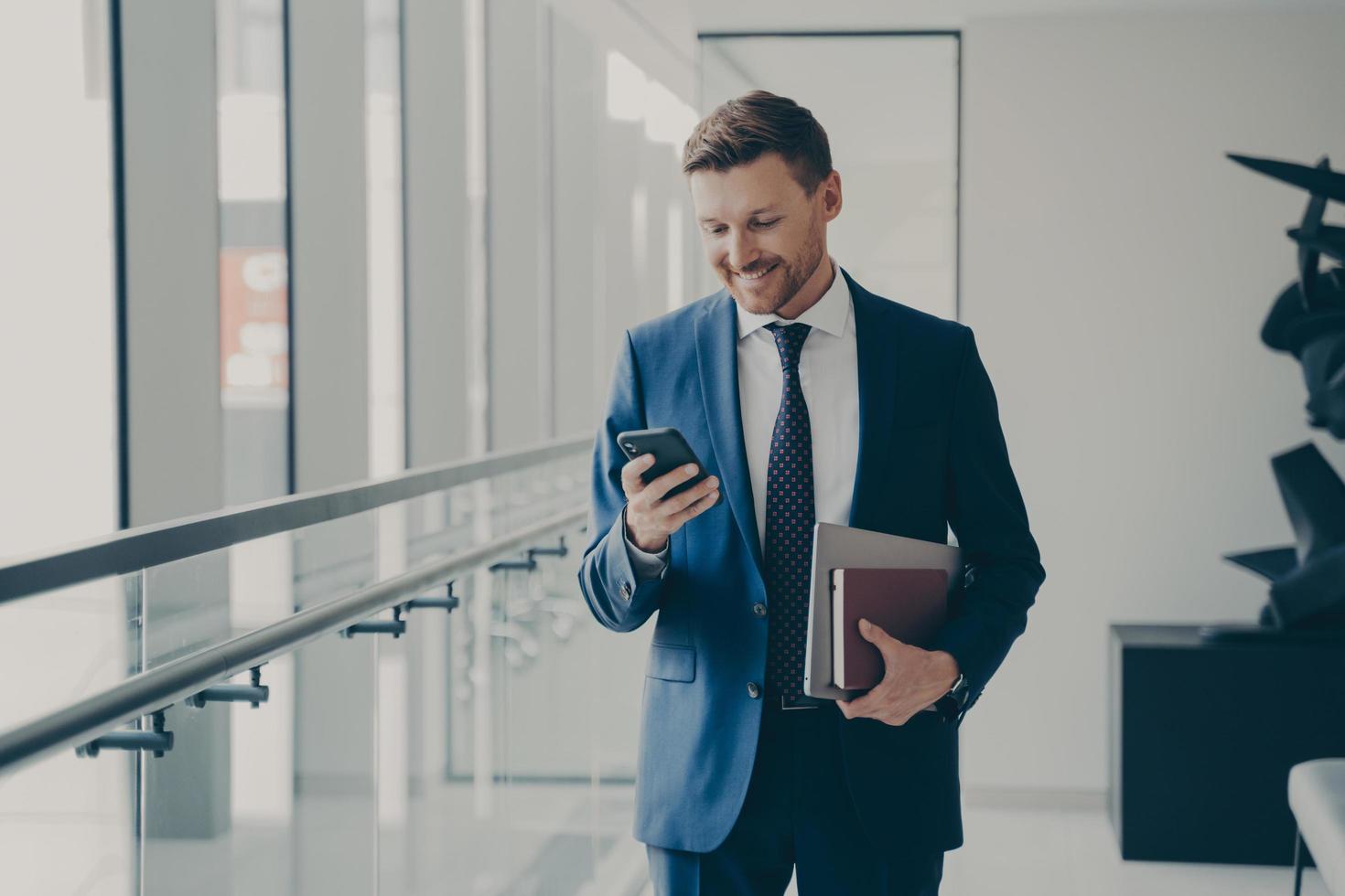 feliz ejecutivo de un hombre guapo sosteniendo un teléfono móvil y sonriendo durante un descanso para tomar café en la oficina foto