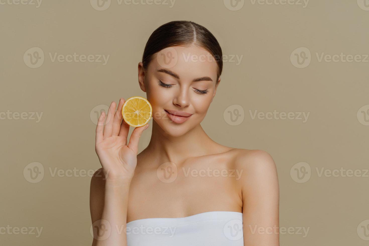 retrato de una encantadora joven morena concentrada sostiene una rodaja de limón fresco recomienda productos de belleza natural prefiere un tratamiento facial orgánico natural envuelto en toallas de baño posa en interiores foto