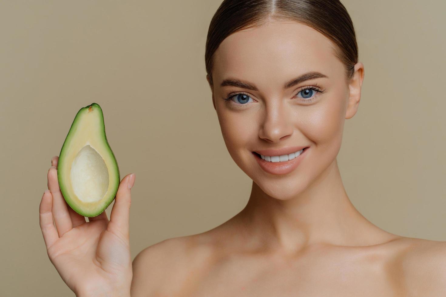 primer plano de una joven modelo caucásica en topless con cabello peinado oscuro sonríe sostiene suavemente la mitad de los soportes de aguacate sin camisa interior se preocupa por la piel y la tez. concepto de bienestar de belleza foto