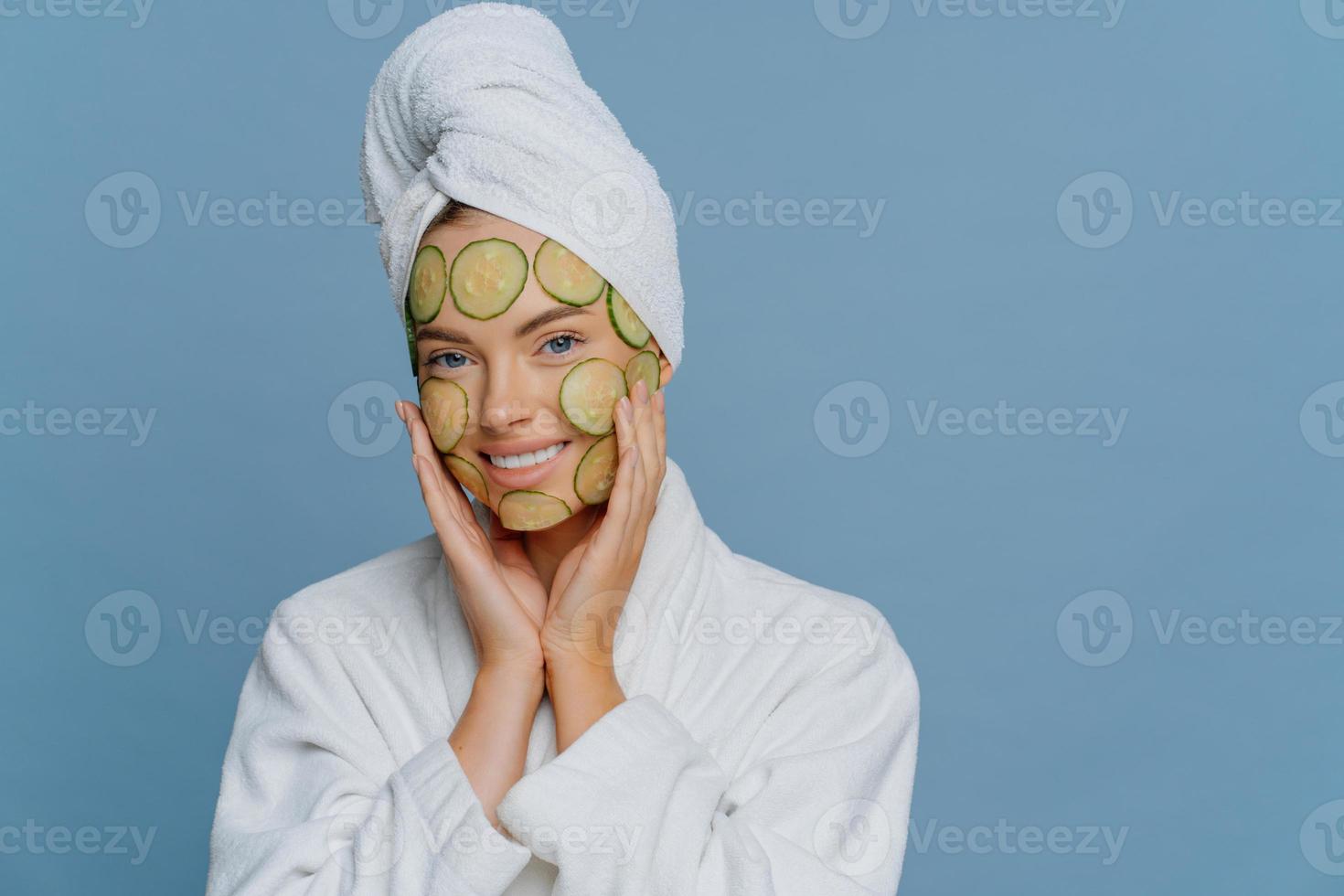 Skin care and beauty treatment concept. Happy relaxed young female model applies cucumber slices to nourish skin keeps hands on face dressed in white dressing gown and wrapped towel on head. photo