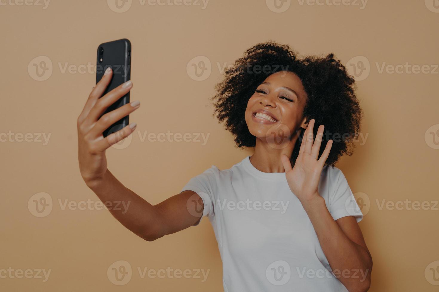 Positive happy mixed race woman waving with hand at camera during video call on modern smartphone photo