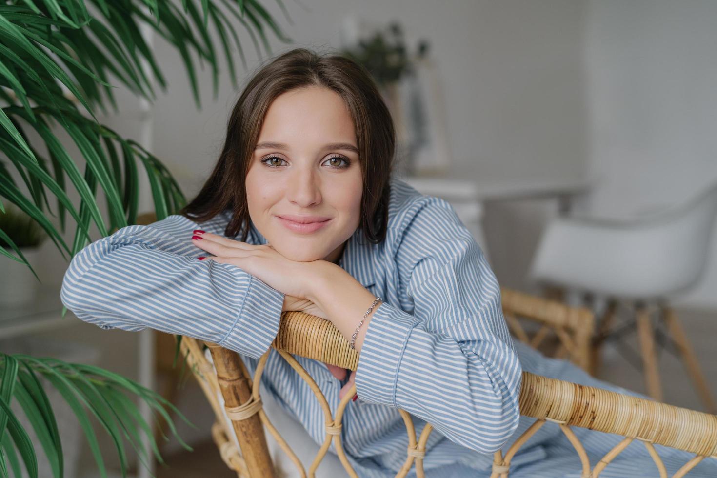 toma horizontal de una atractiva joven caucásica tranquila que tiene una expresión facial complacida, disfruta de un ambiente doméstico, posa en muebles de mimbre, cerca de una planta verde. concepto de personas, belleza y descanso foto