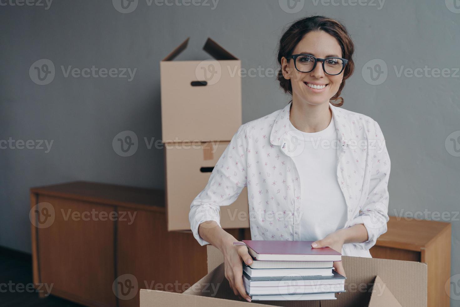 el nerd feliz con gafas está apilando libros en cajas. joven española embalando un contenedor de cartón. foto