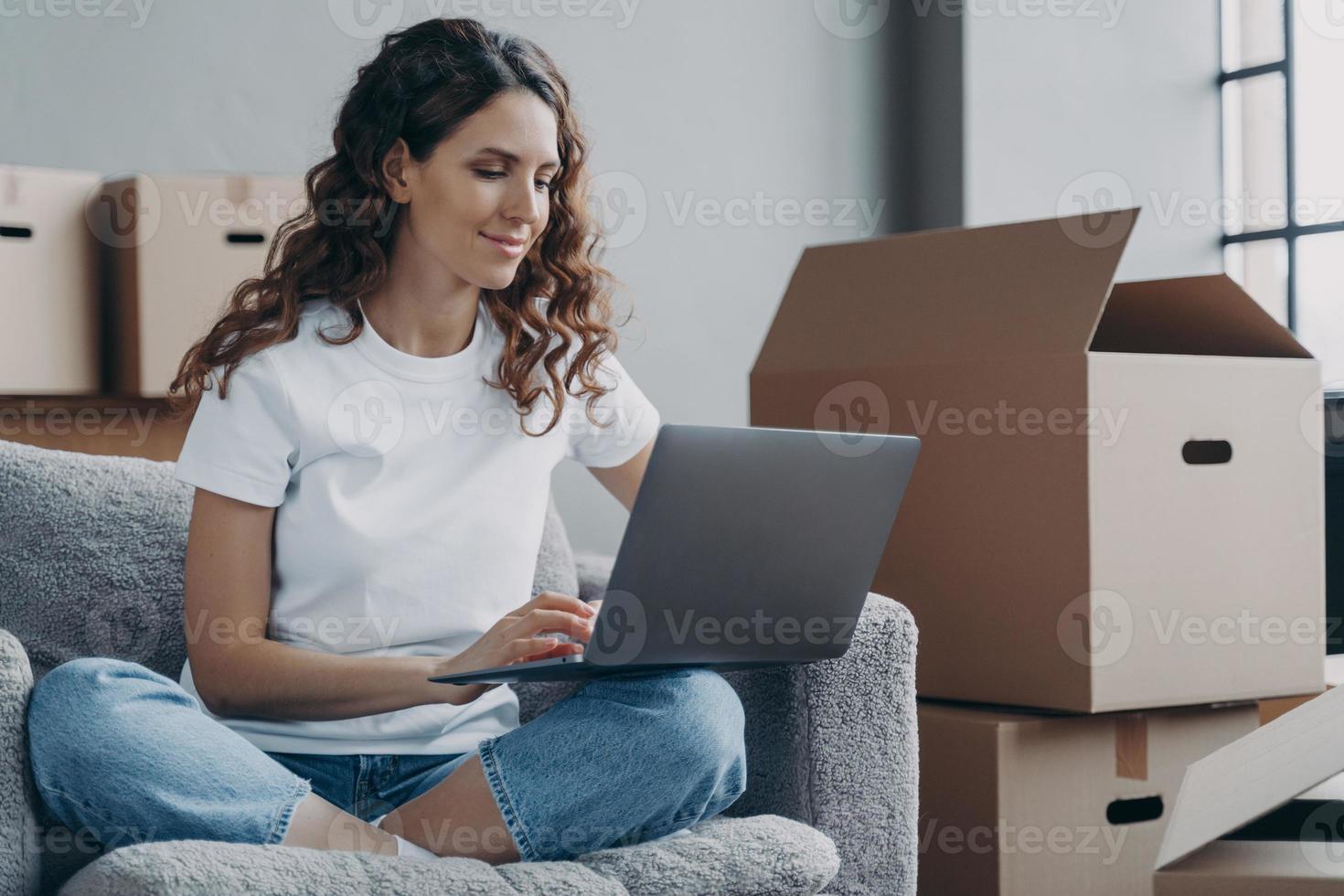 chica hispana sentada con laptop entre cajas de cartón y sonriendo. reubicación y trabajo en línea. foto