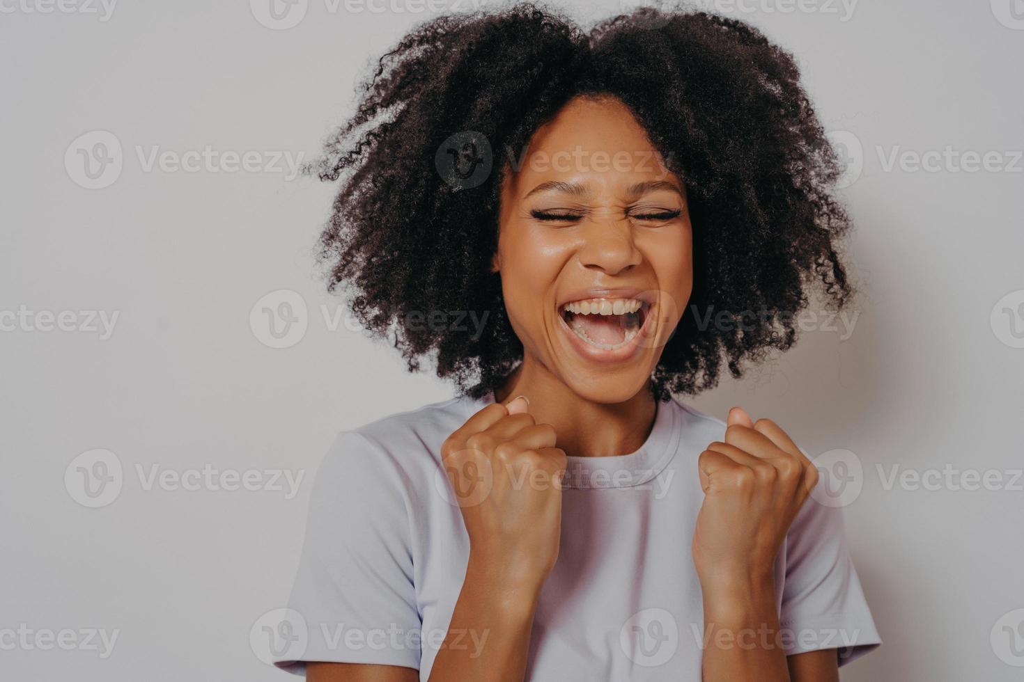 emocionada alegre chica de piel oscura aprieta los puños mientras cierra los ojos, exclama con victoria foto