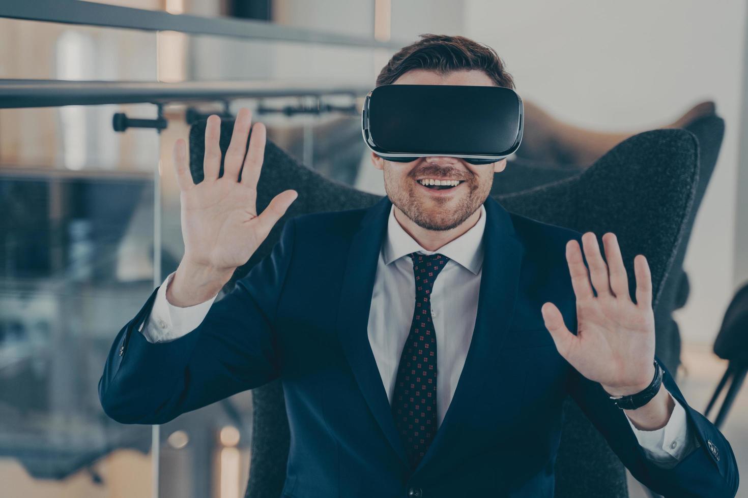 Young smiling businessman in formal black suit sitting in office interior using VR headset photo